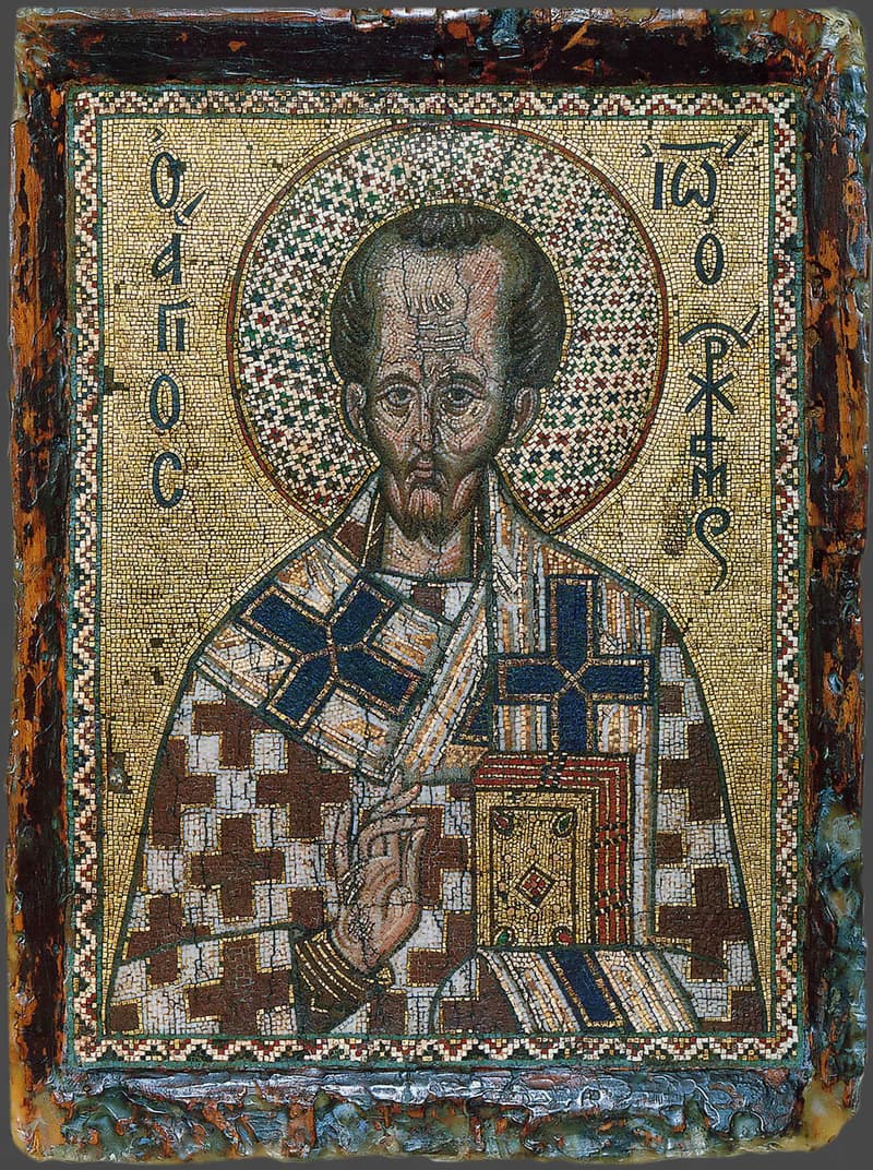 Mosaic icon of St. John Chrysostom, a gaunt, high-browed bishop with a short beard and a patterned gold halo, wearing a white stole marked with crosses, raising his right hand in blessing and holding a jeweled Gospel book, with his name in Greek letters on a gold ground.