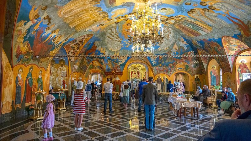 Interior of an Orthodox cathedral during the Divine Liturgy, with frescoes, candles, and worshippers gathered before the iconostasis