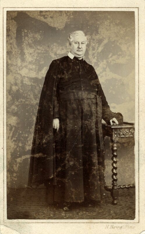 Frederick William Faber, the Anglican convert to Catholicism who wrote the hymn 'Faith of Our Fathers', photographed standing in a long clerical cassock and cloak beside a carved side table