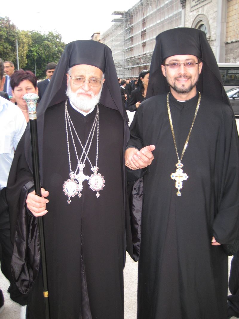Two Melkite Greek Catholic clergy in black ryasa cassocks and bishop's klobuk, wearing pectoral crosses, standing outside a church
