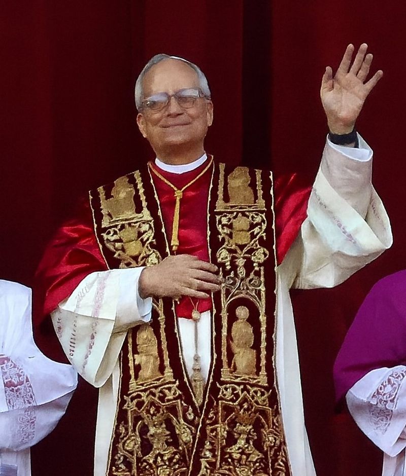 Pope Leo XIV in white papal vestments on the central benediction loggia of St. Peter's Basilica, waving to the crowd during his first public appearance after his election.