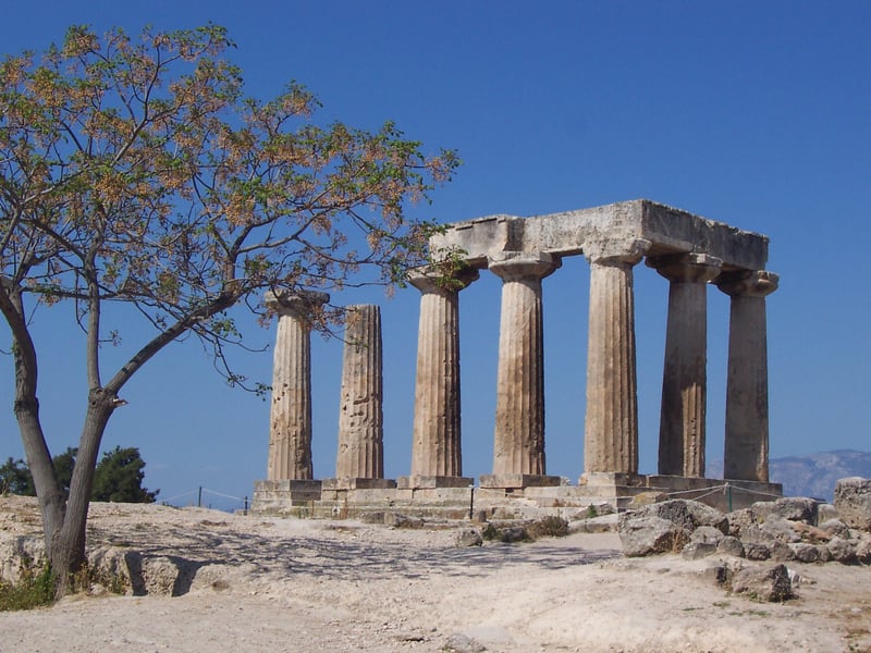 The surviving Doric columns of the mid-6th-century BC Temple of Apollo at Ancient Corinth, standing on a rocky outcrop with Acrocorinth rising in the background