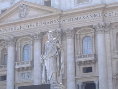 Marble statue of St. Paul holding a sword and book, set against the Renaissance facade of St. Peter's Basilica in Rome.