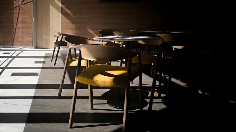 Empty restaurant chairs and tables half-lit by sunlight falling through a window, the other half receding into shadow