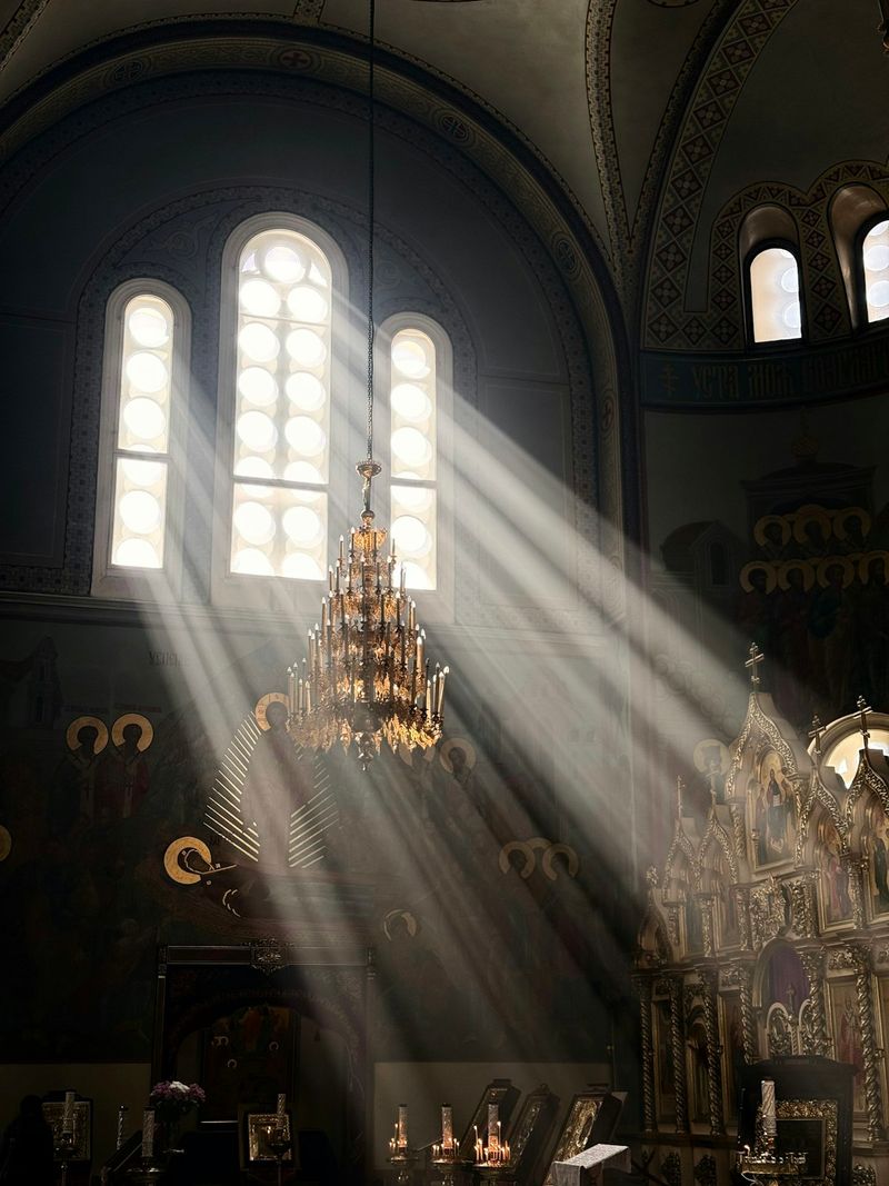 Sunbeams streaming through arched windows of an Orthodox church, illuminating the chandelier and iconostasis
