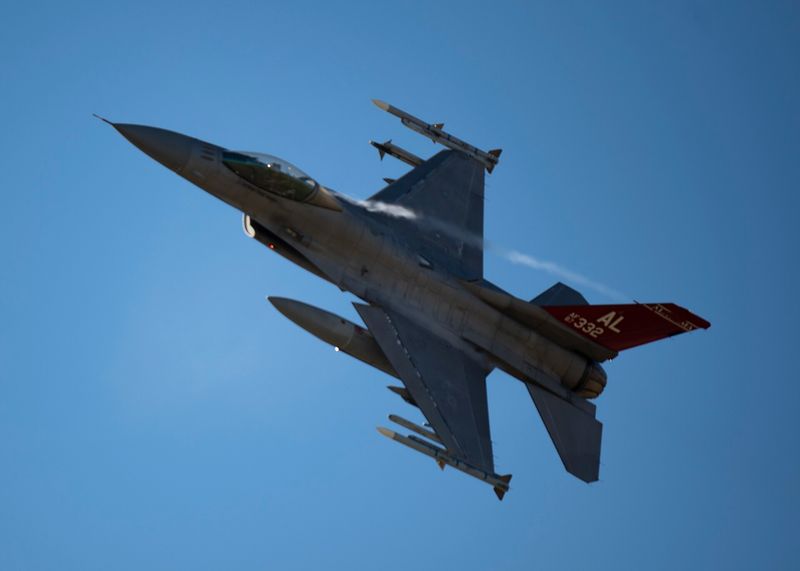 An F-16 Fighting Falcon banking against a blue sky, photographed from below, with a red Alabama tail flash, air-to-air missiles on the wingtips, and condensation streaming from the airframe